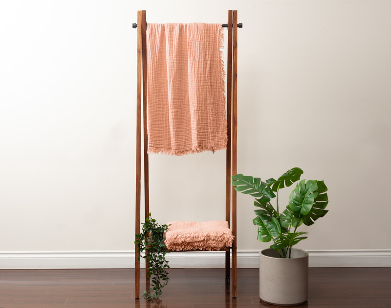 Front view of our Muslin Gauze Throw in Clay hanging on a tall fixture next to a short leafy plant.