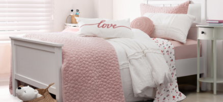 Angled view of our white Lovely Layers collection dressed over a white wooden bed, covered with textured pink quilts and cushions and topped with a pillow with 