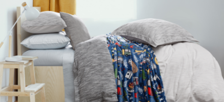 Side view of our grey Modern Mountaineer collection dressed over a twin bed, featuring grey and white covers and a blue fleece blanket decorated with pictures of trees & camping equipment.