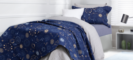 Angled view of our Astro Adventurer collection, dressed over a twin bed in the corner of a white room and featuring a blue quilt covered in constellations and stars, layered just above a checkered grey duvet cover.