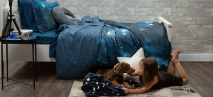 Side view of a young girl playing with a dog on the floor, next to a bed dressed with our Supernova space duvet cover and plush Star and Moon cushions.