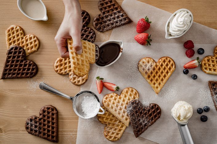 A person dips a heart shaped waffle into a pot of chocolate sauce.