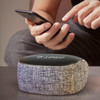 A Cylon Bluetooth speaker in grey fabric with a black top, beside a hand holding a smartphone.