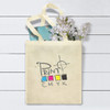 A natural canvas tote bag in cream, featuring a logo and filled with flowers and a blue item.