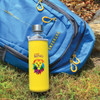 A glass drink bottle in a yellow neoprene sleeve with a lion design, placed next to a blue backpack.