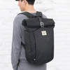 A black roll top backpack is worn by a person against a light brick wall. The backpack features a logo.