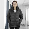 A woman wearing a black puffer jacket with a hood, standing in a modern indoor setting. The jacket has a zip closure.
