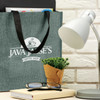 A grey tote bag with black handles featuring a logo, placed beside a lamp, glasses, and a potted plant.