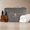 A grey cosmetic bag sits beside two amber bottles and a rolled white towel, featuring a logo on the front.