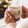 A coffee mug featuring a logo with green leaves and oranges, with a frosted finish and grey base, held in hands.
