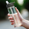 A drink bottle with a clear body, black lid, and straw, held in a hand outdoors.