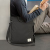 A black crossbody bag rests against a wall with a person sitting beside it, holding books. The bag features a logo.