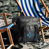 A grey camo cooler bag sits next to striped beach chairs on sand, featuring a logo.