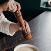 A wooden pepper mill being operated by a person, with a bowl of creamy mixture on a dark surface.