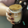 A drink cup made of glass with a bamboo lid, featuring a logo and filled with a layered coffee beverage.