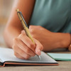 A wooden pen with a sleek gold finish, being held while writing in a notebook. The pen features a branding logo.
