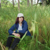 A woman sits in tall grass wearing a navy soft shell jacket and a light-coloured cap, surrounded by trees.
