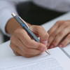 A metal pen in grey is being held, with a logo visible. A hand is writing on a sheet of paper.