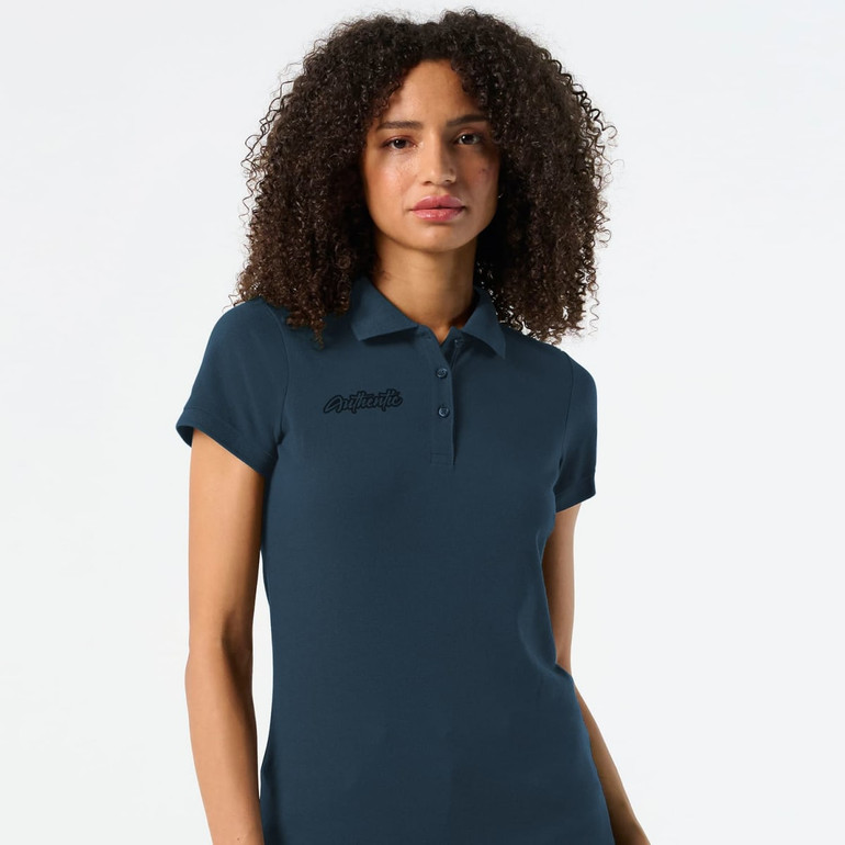 A dark teal women's polo shirt with a slim fit and a logo on the chest. The model has curly hair and wears the shirt.