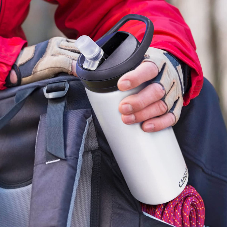 A drink bottle in silver with a black lid, held by a person in gloves, resting against a bag.