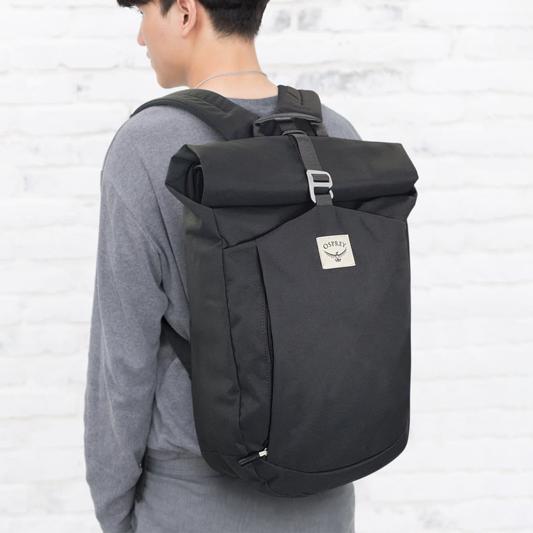 A black roll top backpack is worn by a person against a light brick wall. The backpack features a logo.