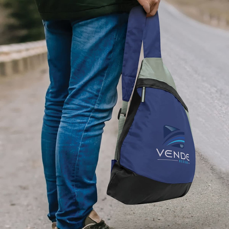 A slinger bag in navy blue with grey and black accents, featuring a logo. It is being held by a person on a path.