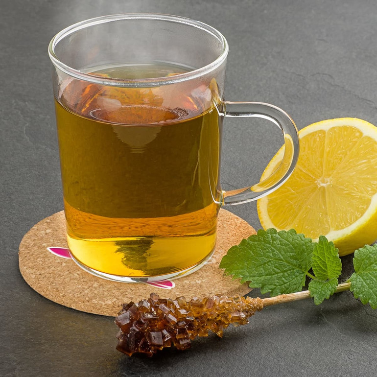 A round oak cork coaster sits beneath a glass mug of herbal tea, alongside a lemon slice and a sugar stick.