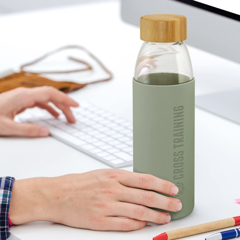 A drink bottle with a glass body, green silicone sleeve, and bamboo lid, resting on a desk next to a computer.