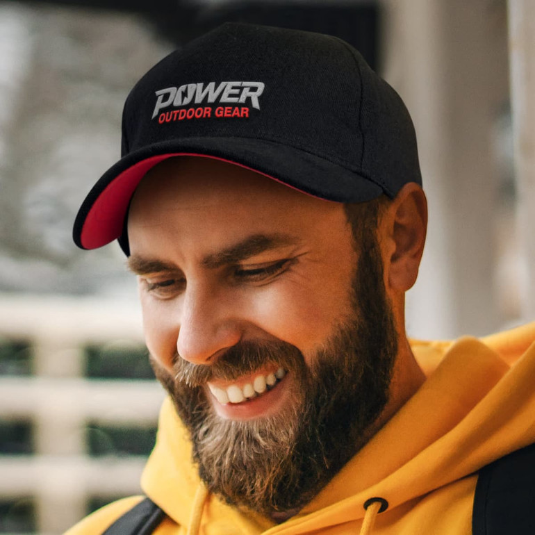 A black and red cap featuring a logo, worn by a smiling man with a beard, against an outdoor backdrop.