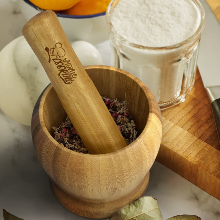 A bamboo mortar and pestle set, featuring a wooden pestle and herbs inside the mortar, with a glass jar and granite surface nearby.