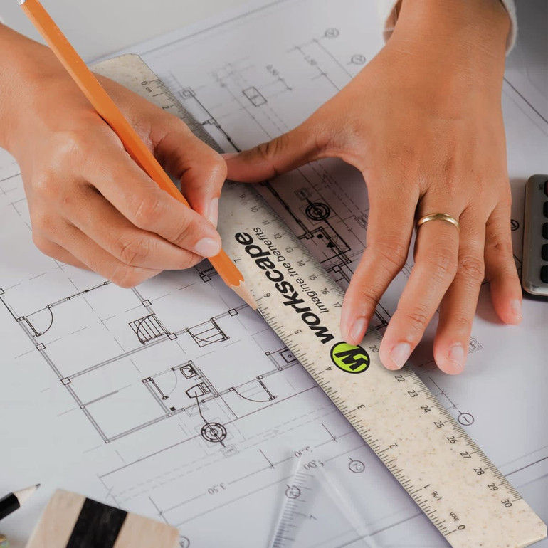 A 30cm ruler with a logo, being held over architectural plans while a hand uses a pencil. The ruler is a transparent colour with metal edges.