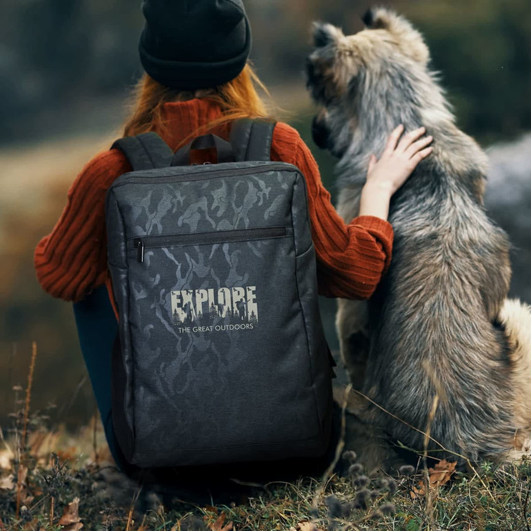 A grey Urban Camo Backpack rests against a person in a red sweater, next to a fluffy dog in a natural outdoor setting.