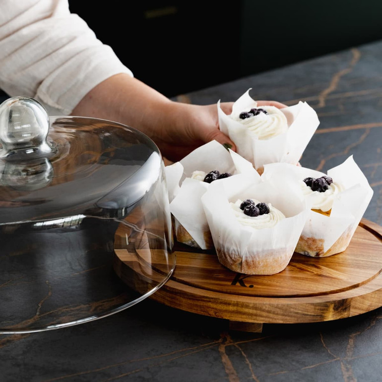 A wooden cake display holding cupcakes, covered with a clear glass dome, featuring white frosting and blueberries.