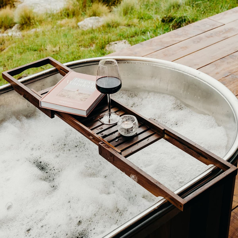 A wooden bath caddy in dark brown, holding a glass of red wine, a candle, and a book, resting over a bubbly bathtub.
