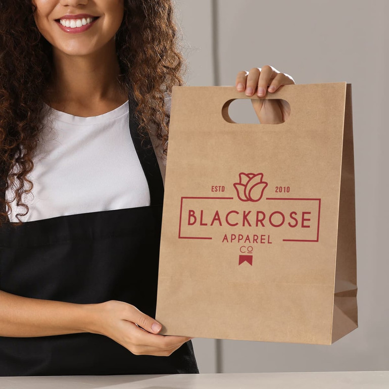 A woman holds a large, brown die cut paper bag with a decorative logo on the front.