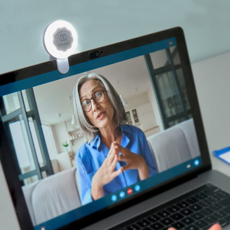 A white Halo Selfie Light attached to a laptop, illuminating a person on a video call. The scene features a modern interior.