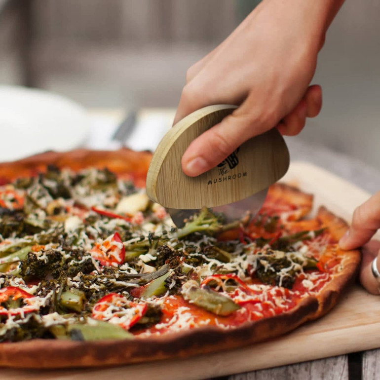 A bamboo pizza cutter with a rounded handle, slicing through a vegetable-topped pizza.