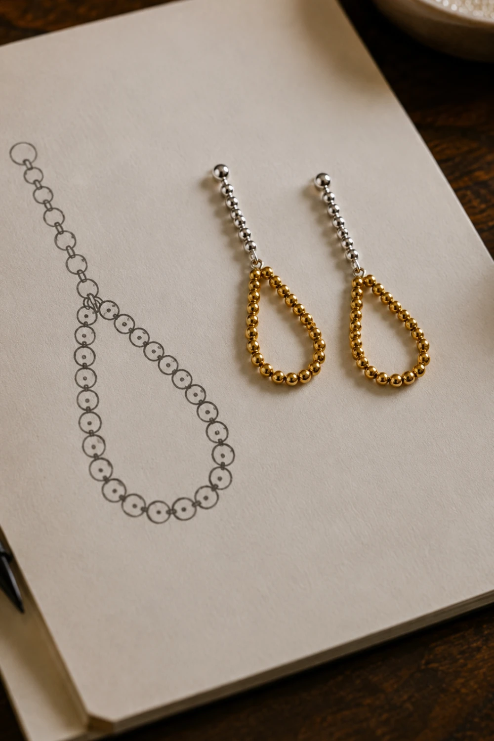 Silver and gold drop earrings beside an earring design sketch on cream paper