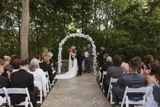 A bride and groom say "I do" under a curved arch adorned with white flowers at a wedding ceremony while guests are seated and watch.