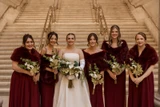 A bride and her bridesmaids wearing maroon dresses and matching maroon fur shawls pose for a photo holding classic green and white bouquets.