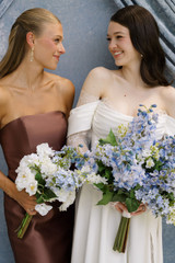 Bride in an off-the-shoulder white wedding gown stands beside a bridesmaid in a mocha satin dress, each holding blue and white silk bouquets from the Millie Collection featuring hydrangeas, delphinium, white garden roses, and lush greenery. The pair smile at one another against a soft blue backdrop.