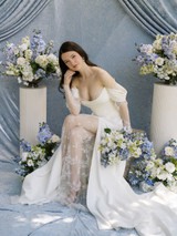 Bride in an off-the-shoulder white lace wedding gown sits surrounded by lush Millie Collection silk floral arrangements featuring blue hydrangeas, delphinium, white garden roses, and fresh greenery. The editorial bridal portrait is styled against a soft blue draped backdrop with coordinating floral meadows and elevated pedestal arrangements.