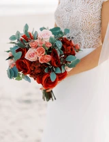A bride holds her lush red and light pink bridal bouquet close to her as she poses on the beach for a photo.
