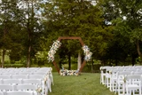 A wooden hexagon arch is adorned with multi-colored pastel floral runners for a lush ceremony backdrop installation in an outdoor setting with white chairs for guest seating.