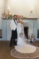 A bride and groom stand in front of a light blue tiled fire place with light pink, ivory, and light green florals sit on top of the mantle and at the base of the fireplace, there are coordinating light pink, ivory, and light green florals.