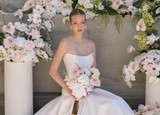 Bride in a modern white gown sits among lush floral installations featuring blush anthuriums, white orchids, pale pink roses, and soft green hydrangeas while holding a bouquet of blush and ivory blooms.