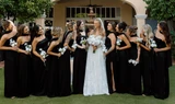 A bride and her bridesmaids wearing black dresses holding white rose and hydrangea bouquets outside in front of an elegant wedding venue