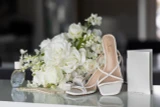 White bridal heels styled with white rose arrangements, wedding rings, and bridal accessories on a reflective surface.”