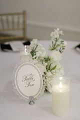 White floral arrangement placed in the center of a round table next to a table number sign and pillar glass pillar.