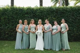 A bride and her bridesmaids wearing sage green dresses holding white rose and hydrangea bouquets outside in front of an elegant wedding venue